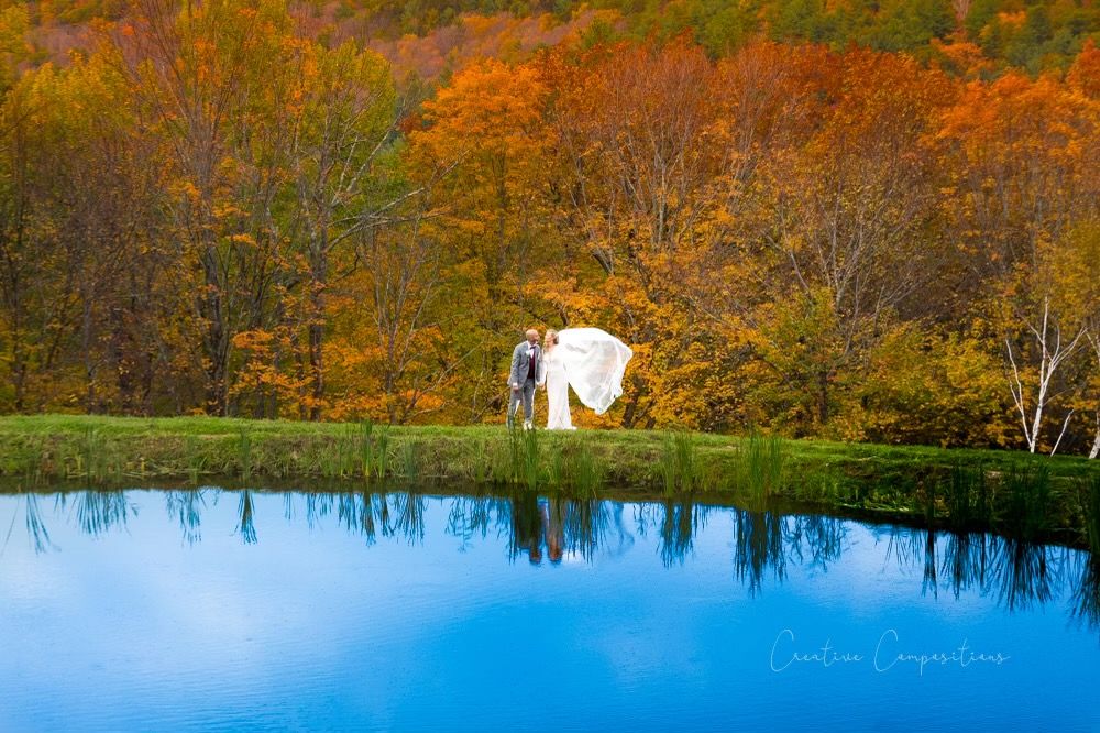 A married couple standing by a reflective pond in the autumn while the bride's veil flows in the wind.